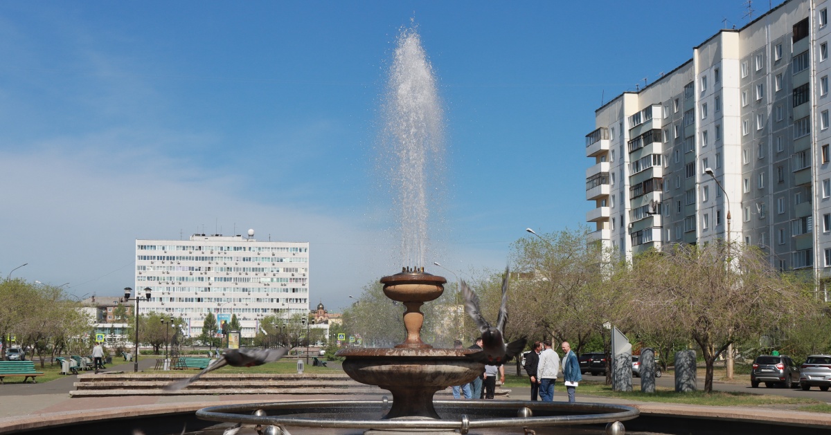 Фонтан в городском парке Братска, окруженный людьми на фоне многоэтажек в солнечный день.