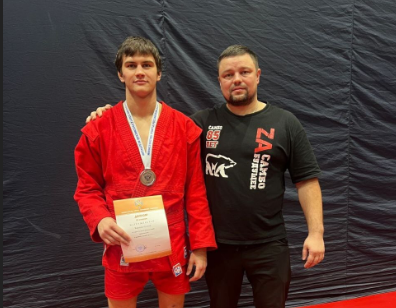 Спортсмен с медалью и тренером на фоне стенда.