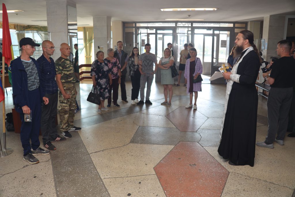 Митинг в помещении, выступает священник, люди слушают.