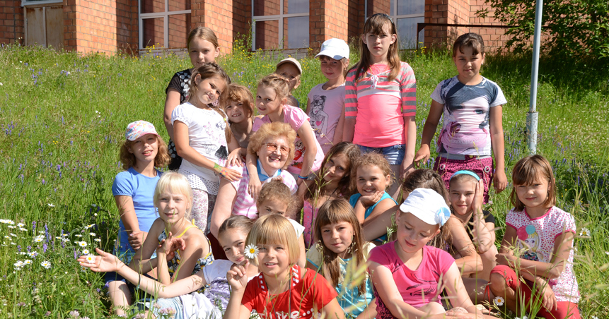 Группа улыбающихся детей на природе перед кирпичным зданием в солнечный день.