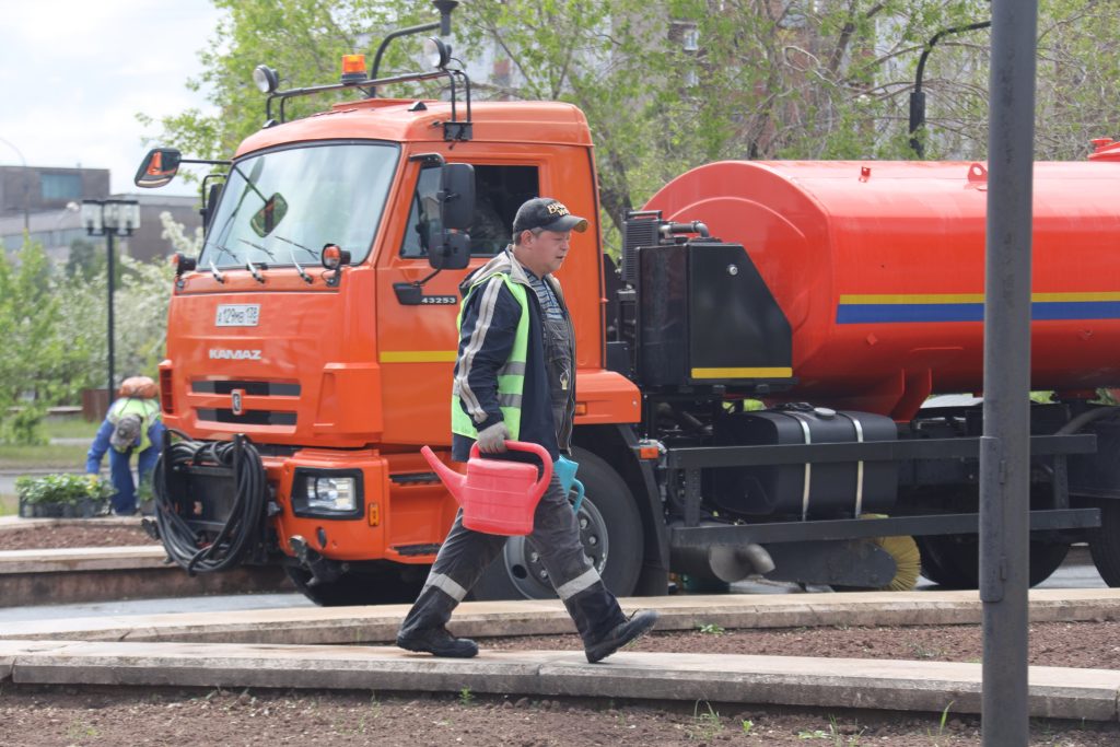 Рабочий с лейкой, оранжевый водовозный грузовик на заднем плане.
